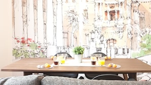 A dining table set with breakfast items, a potted plant, and a mural of architectural details. - Del Parque Flats Santos Catedral - Cozy Apt in Málaga's Historic Center, Walk to Attractions (Málaga)