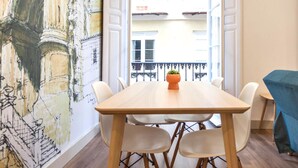 A dining area with a wooden table and white chairs, a potted plant on the table, and a view of buildings through an open door. - Del Parque Flats Santos Catedral - Cozy Apt in Málaga's Historic Center, Walk to Attractions (Málaga)