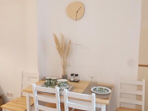Comedor con mesa de madera, sillas blancas, un reloj en la pared y un jarrón con plantas secas.