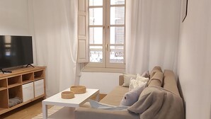 Ett vardagsrum med en tv på ett träställ, en soffa, ett soffbord och ett fönster med gardiner.