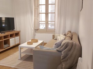 Una sala de estar con sofá, una mesa de centro, un televisor sobre un mueble de madera y una ventana con cortinas.