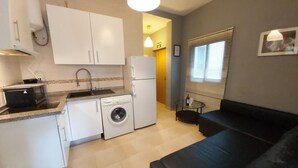 A compact living space with a kitchenette, a washing machine, a refrigerator, a black sofa, a small table with a bottle, and a window with blinds.