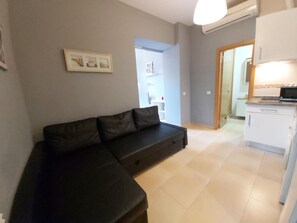 A modern living room with a black leather sofa, a kitchen area with white cabinets, and a door leading to another room.