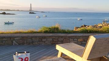 Une terrasse en bois avec une chaise numérotée et un sac, donnant sur une mer calme où l’on aperçoit des bateaux et un phare au loin.