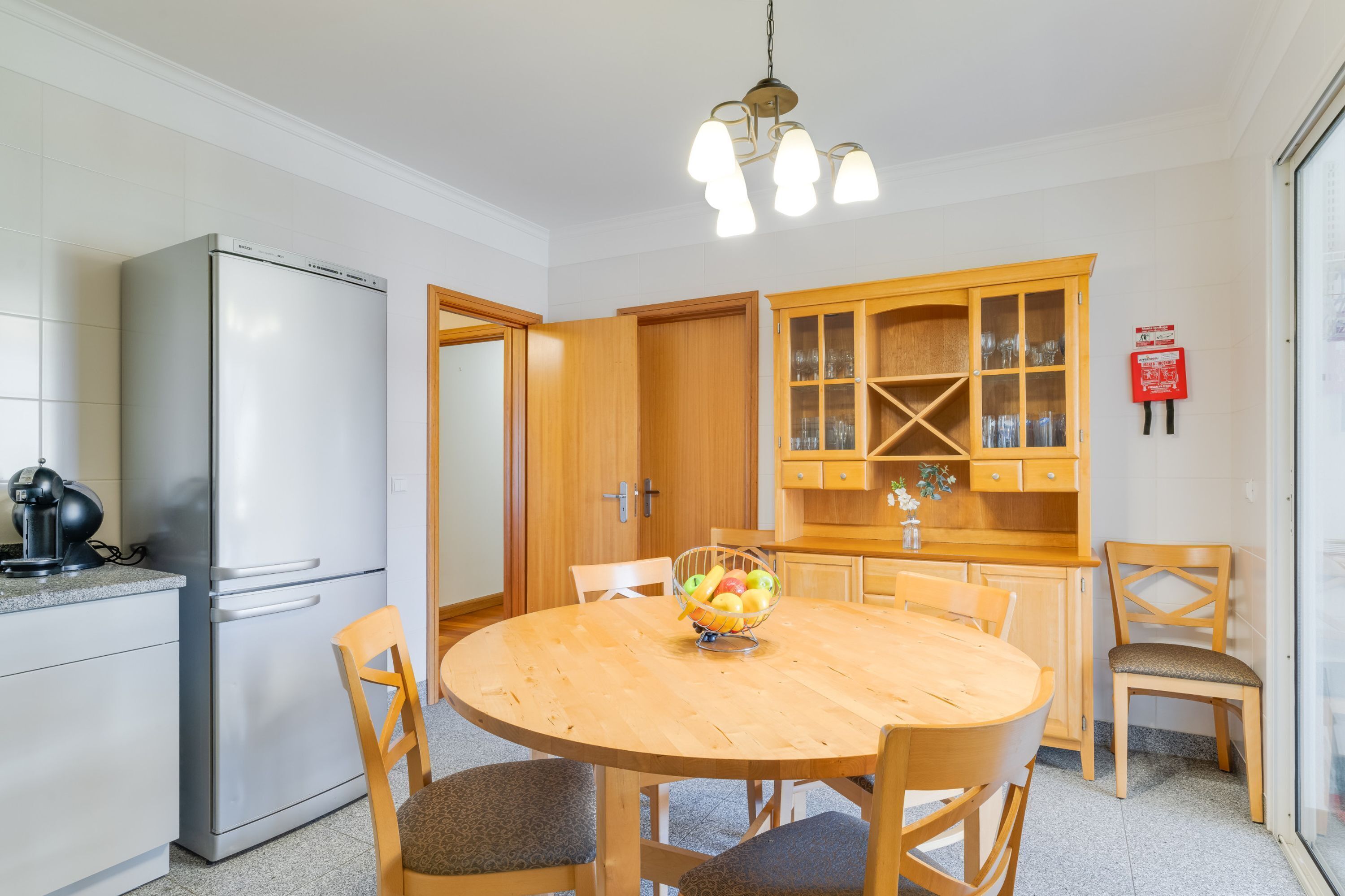 A compact kitchen with a refrigerator, wooden cabinets, a dining table with chairs, and a fruit bowl.