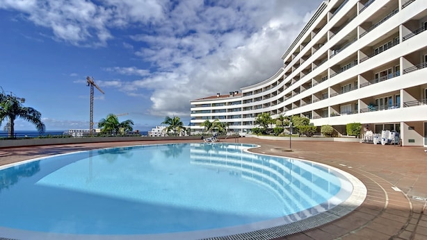 A large outdoor swimming pool surrounded by a multi-story hotel with balconies, palm trees, and a clear sky. - Monumental Ocean View by Atlantic Holiday (Funchal)