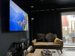 Ein modernes Wohnzimmer mit einem großen Flachbildfernseher an der Wand, einem Sofa, einem Couchtisch und einem kleinen Beistelltisch mit einer Fernbedienung.