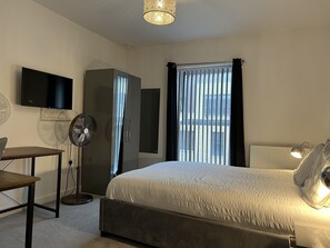 Ein Schlafzimmer mit Bett, Schreibtisch, Fernseher, Kleiderschrank, Standventilator und einem Fenster mit Vorhängen.