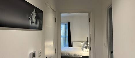 A hallway leading to a bedroom with a bed, a nightstand, and a wall-mounted control panel.