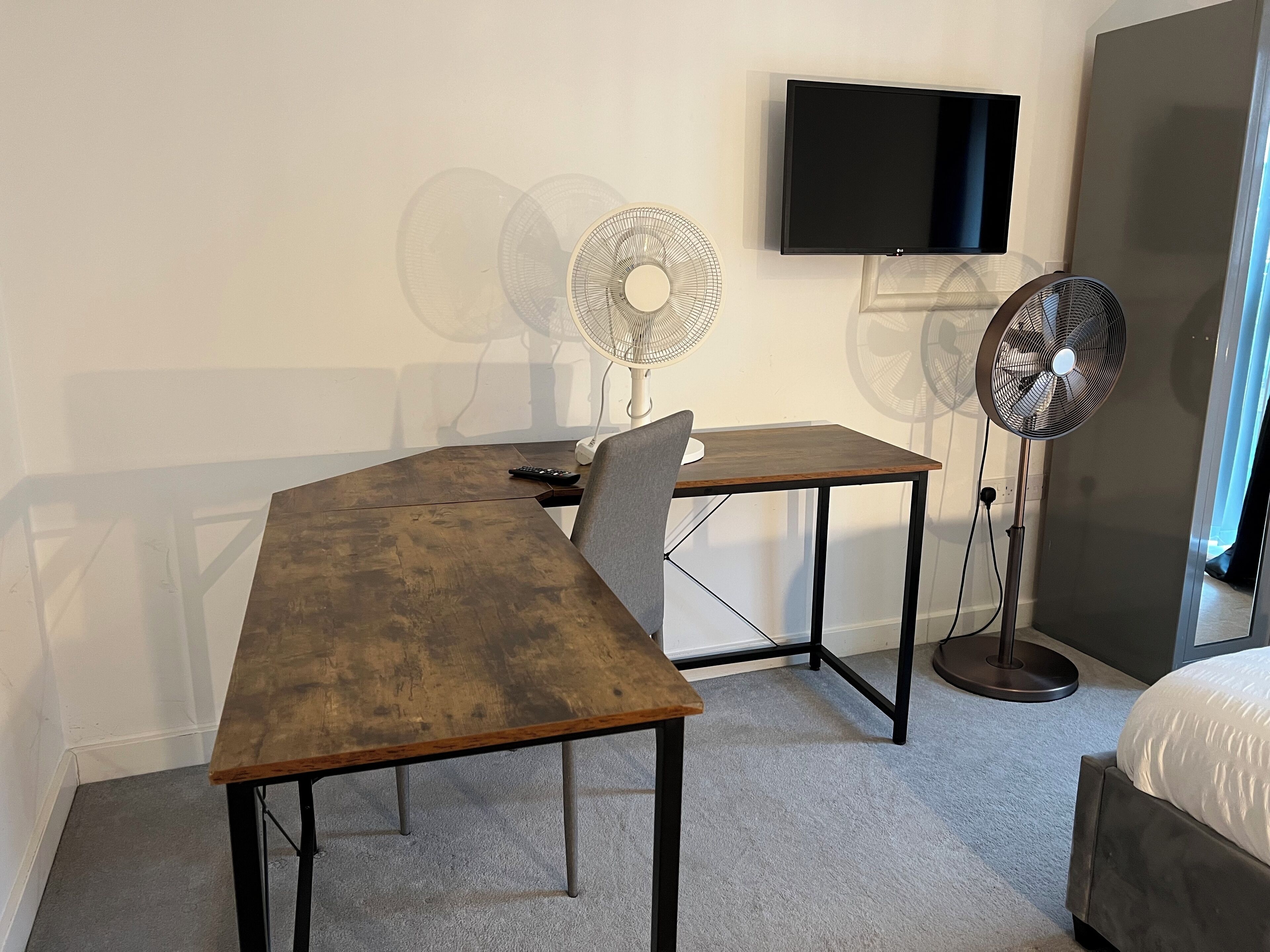 A room with a wooden desk, a chair, a television mounted on the wall, and two standing fans.