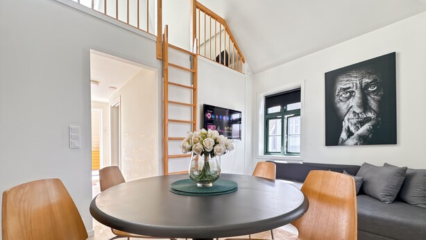 A modern living room with a round table, wooden chairs, a grey sofa, and a large black and white artwork on the wall.