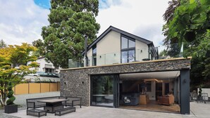 A modern house with a stone facade, large sliding glass doors leading to an open-plan living area with a fireplace, and a patio with outdoor furniture. - Hare's Breath (Windermere)