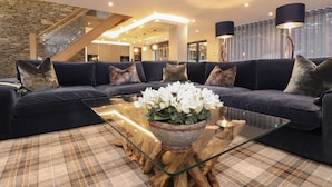 A modern living room with a dark blue sofa, a glass coffee table with a floral centerpiece, and a checkered rug. A stone wall and wooden staircase are visible in the background. - Hare's Breath (Windermere)