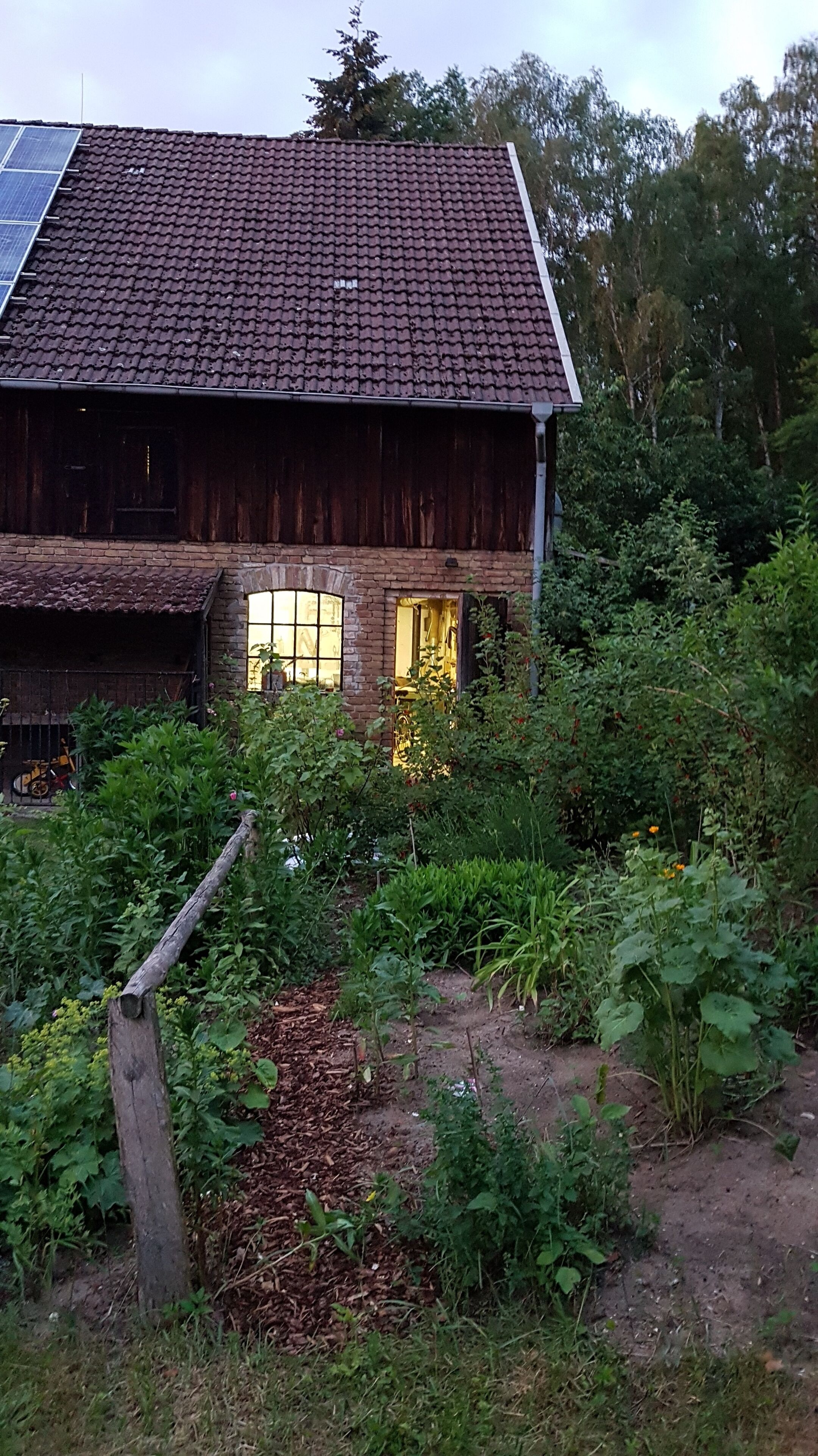 A rustic wooden building with a tiled roof, surrounded by dense greenery and a garden area.