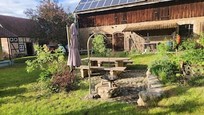 A rustic outdoor area with a wooden table and benches, surrounded by greenery and a stone pathway.