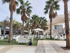 An outdoor seating area with white wicker chairs and tables, surrounded by palm trees.