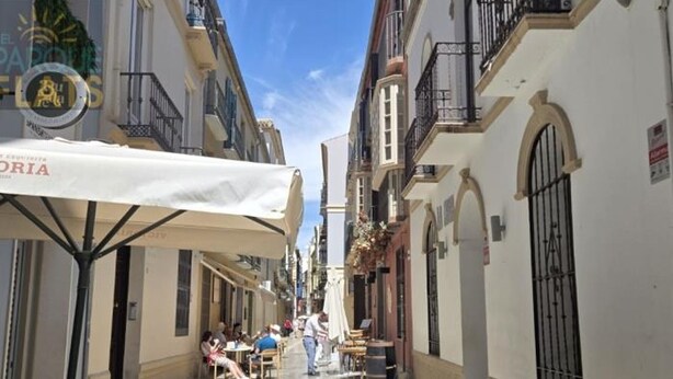 Una calle estrecha con mesas al aire libre, edificios blancos y cielo despejado.