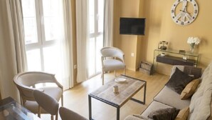 A living room with a sofa, two chairs, a coffee table, a TV, and a clock on the wall. - Del Parque Flats Cervantes: Cozy Apartment for 4 in Historic Málaga Center (Málaga)