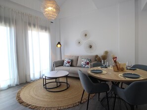 Uma sala de estar moderna com uma mesa de jantar redonda, sofá cinzento e uma grande janela com cortinas leves.