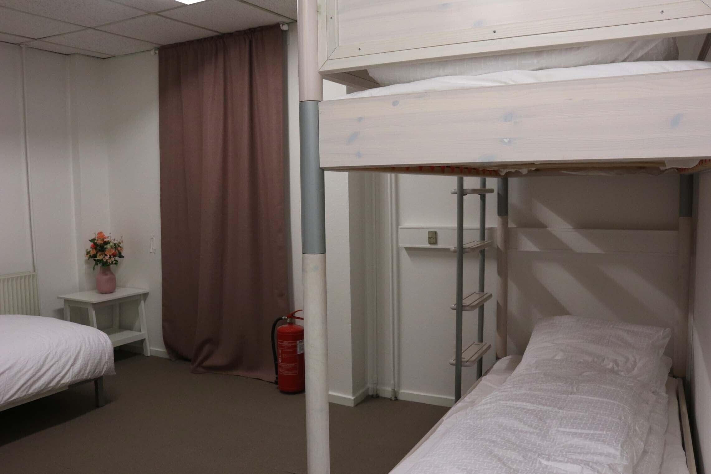 A dormitory room with bunk beds, a small bedside table with a vase of flowers, a fire extinguisher, and a curtain.