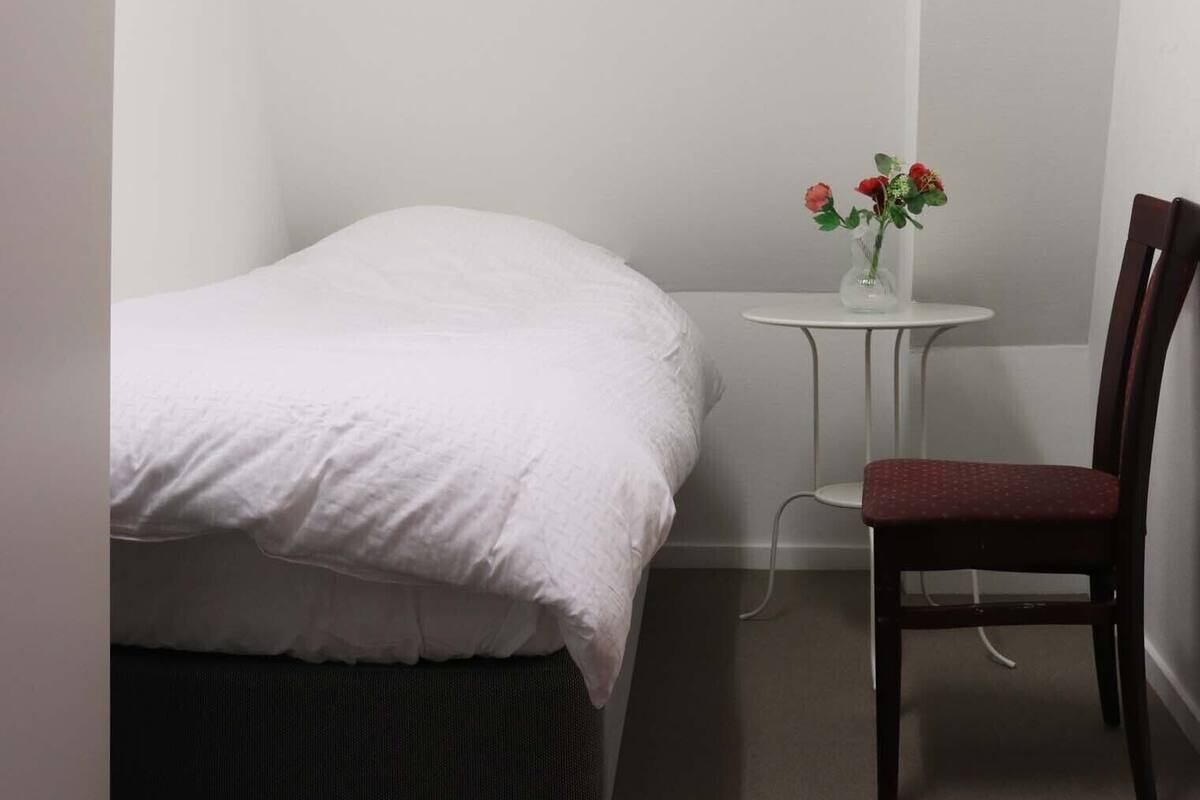 Une petite pièce, bien rangée, avec un lit, une chaise et une petite table sur laquelle est posé un vase de fleurs.