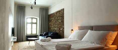 A modern hotel room with a large bed, a stone accent wall, a seating area with a small table, and a large window with curtains.