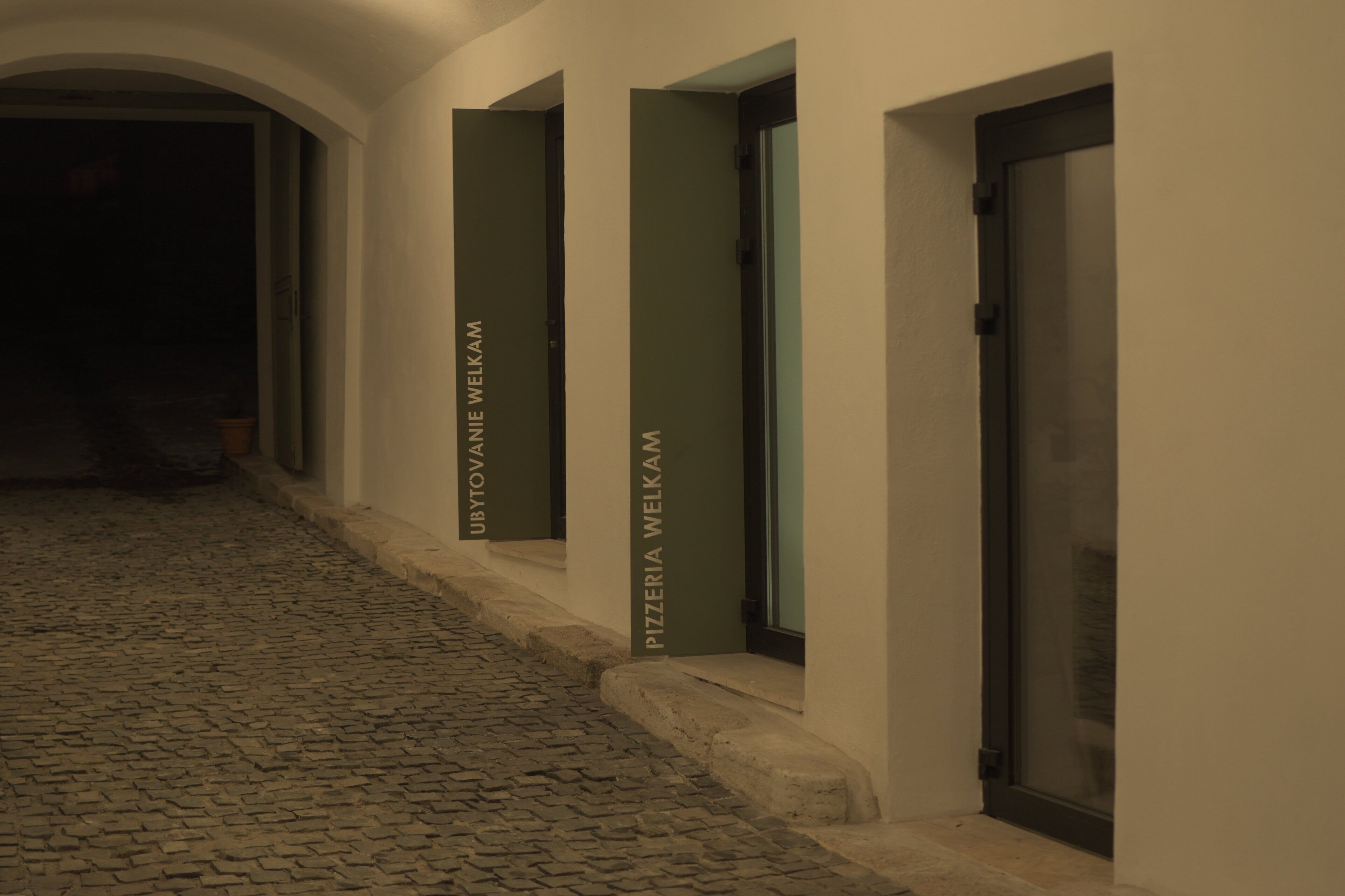 Un pasillo con piso de adoquines y puertas con la leyenda 'UBYTVA ANIE WELKAM' y 'PIZZERIA WELKAM'.