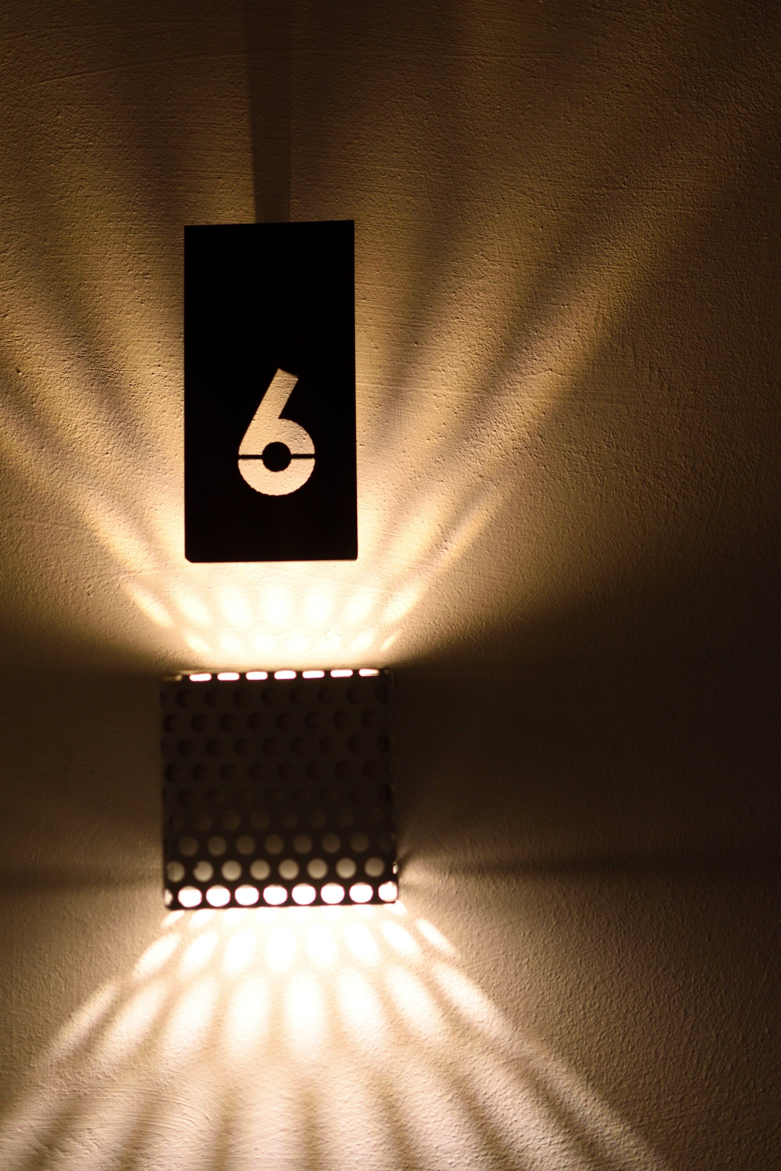 A wall-mounted light fixture with a number '6' sign, casting a warm glow and creating a pattern of light and shadow on the wall.