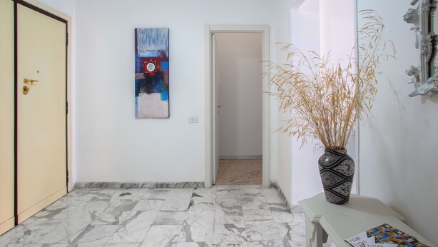 Un pasillo con suelo de mármol, un cuadro en la pared y un jarrón con plantas secas sobre una mesa.