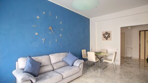 Una sala de estar con pared azul, sofá blanco y comedor con mesa y sillas.
