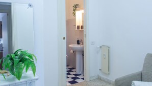 Una habitación con pared blanca, un espejo, una planta verde, un sofá gris y un baño visible a través de una puerta abierta.