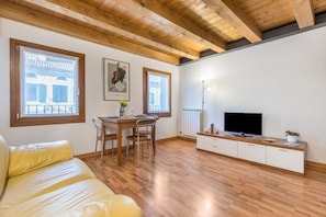 A room with wooden beams, a sofa, a TV, a dining table with chairs, and windows with blinds.