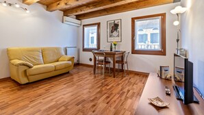 Ein Zimmer mit Holzbalkendecke, einem gelben Sofa, einem Esstisch mit Stühlen und einem Fernseher.