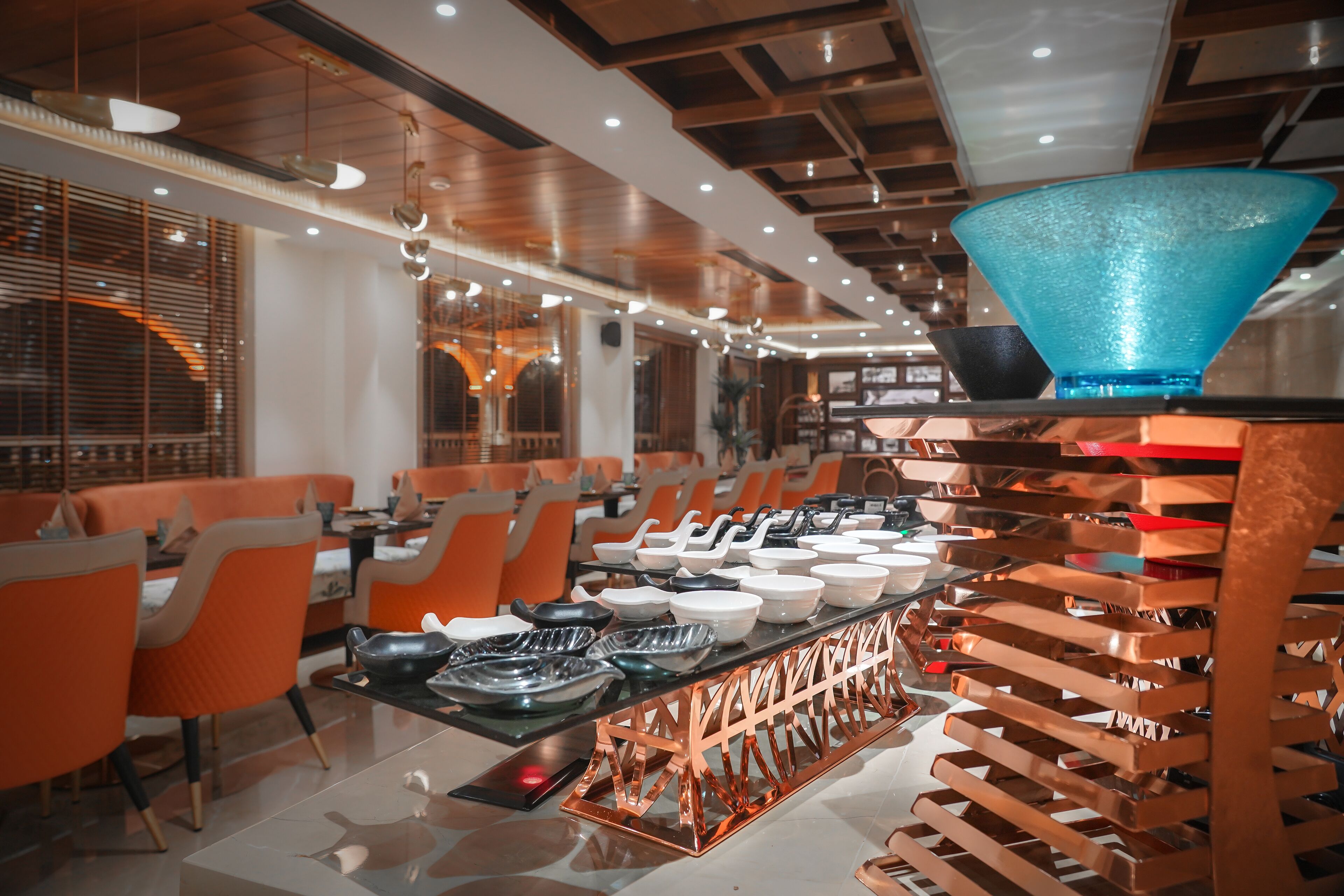 A modern restaurant interior with orange chairs, a central dining table with various dishes, and a unique blue glass bowl on a stand.