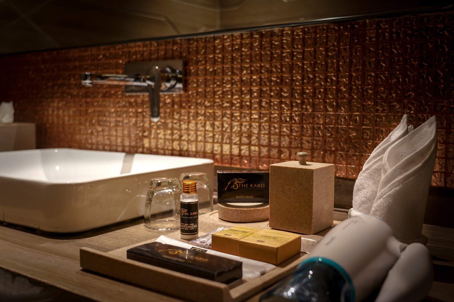A bathroom with a large sink, a mirror, and a textured wall. There are toiletries on the counter, including a bottle of soap, a bar of soap, and a box of tissues.