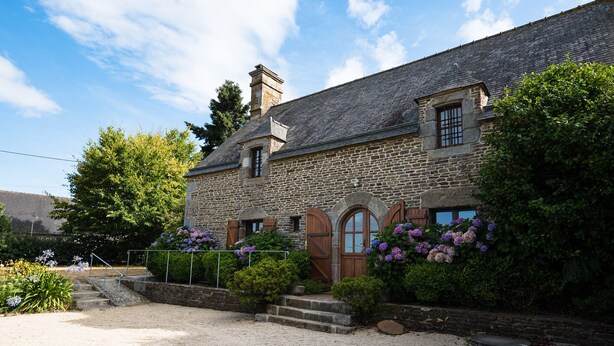 A stone building with a traditional design, featuring a wooden door and window shutters, surrounded by lush greenery and a gravel driveway. - Charming gîte with Spa, on the bay of Mont Saint Michel (Sougeal)