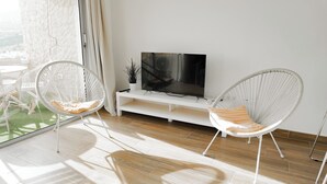 Ett modernt vardagsrum med en platt-TV på ett vitt TV-bord, två vita stolar med stomme av metalltråd och kuddar, samt en stor glasdörr som leder ut till en balkong med utsikt över stadsbilden.