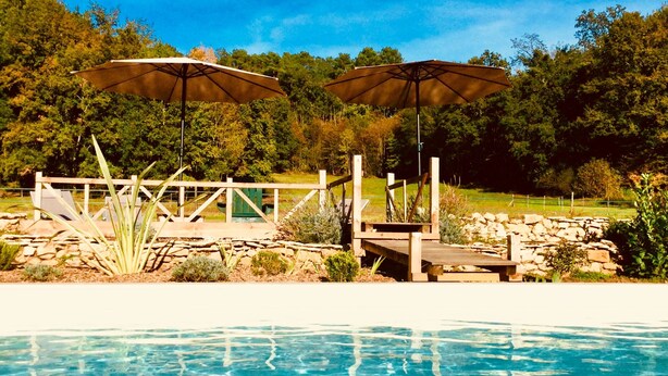 A swimming pool with clear blue water, surrounded by a wooden deck and two umbrellas.
