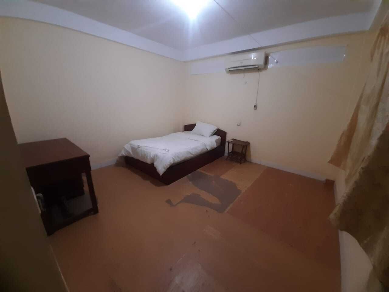 A small, single-bed room with a brown floor, a bedside table, and an air conditioning unit mounted on the wall.