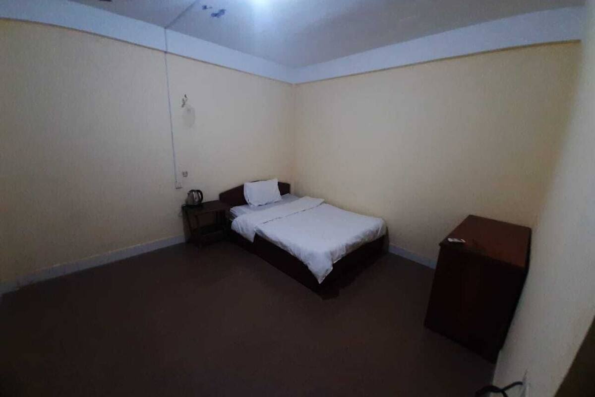 Een kleine, zwak verlichte kamer met een enkel bed, een nachtkastje en een houten kast.