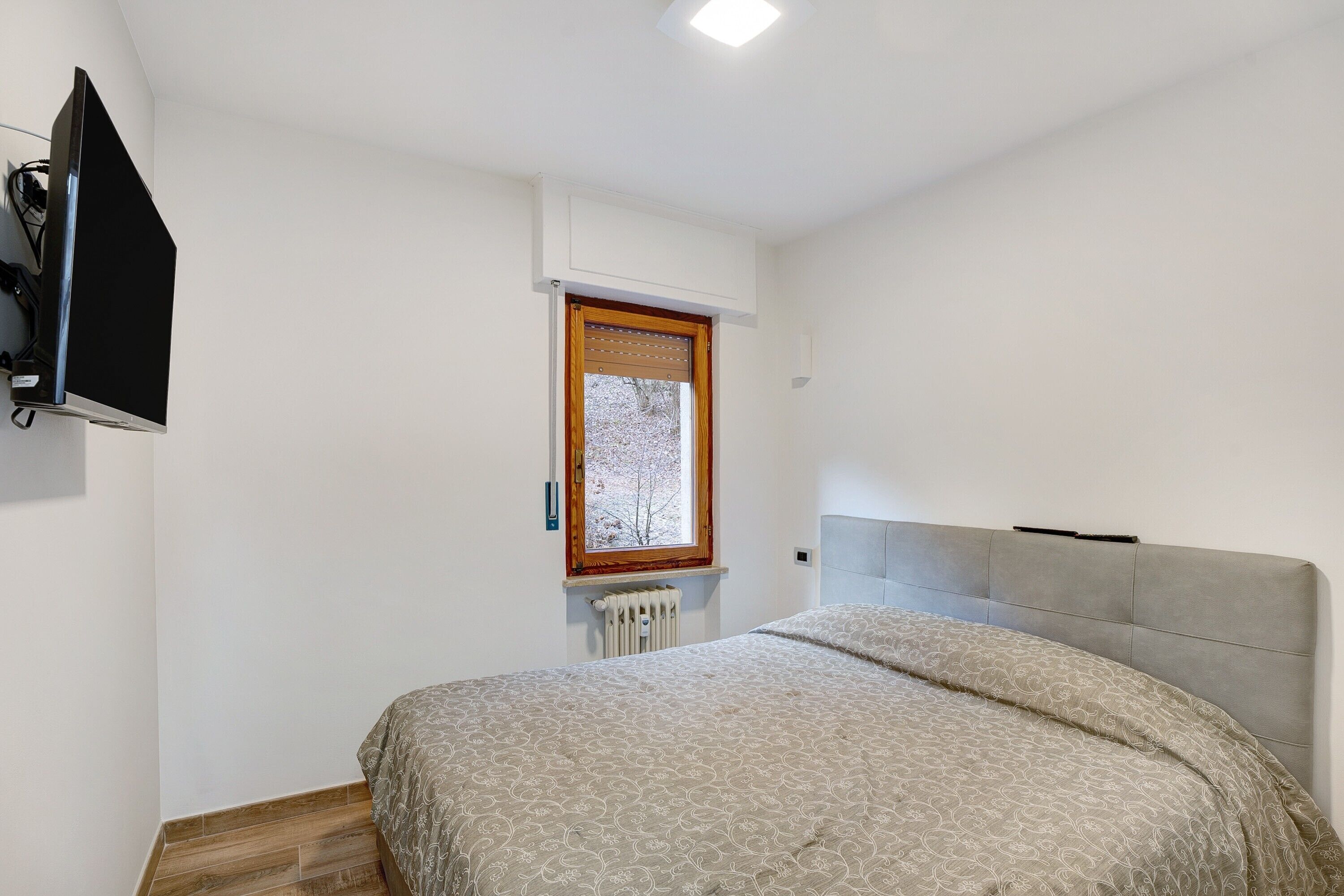 Ein Schlafzimmer mit Bett, Fenster, Heizkörper und einem an der Wand montierten Fernseher.