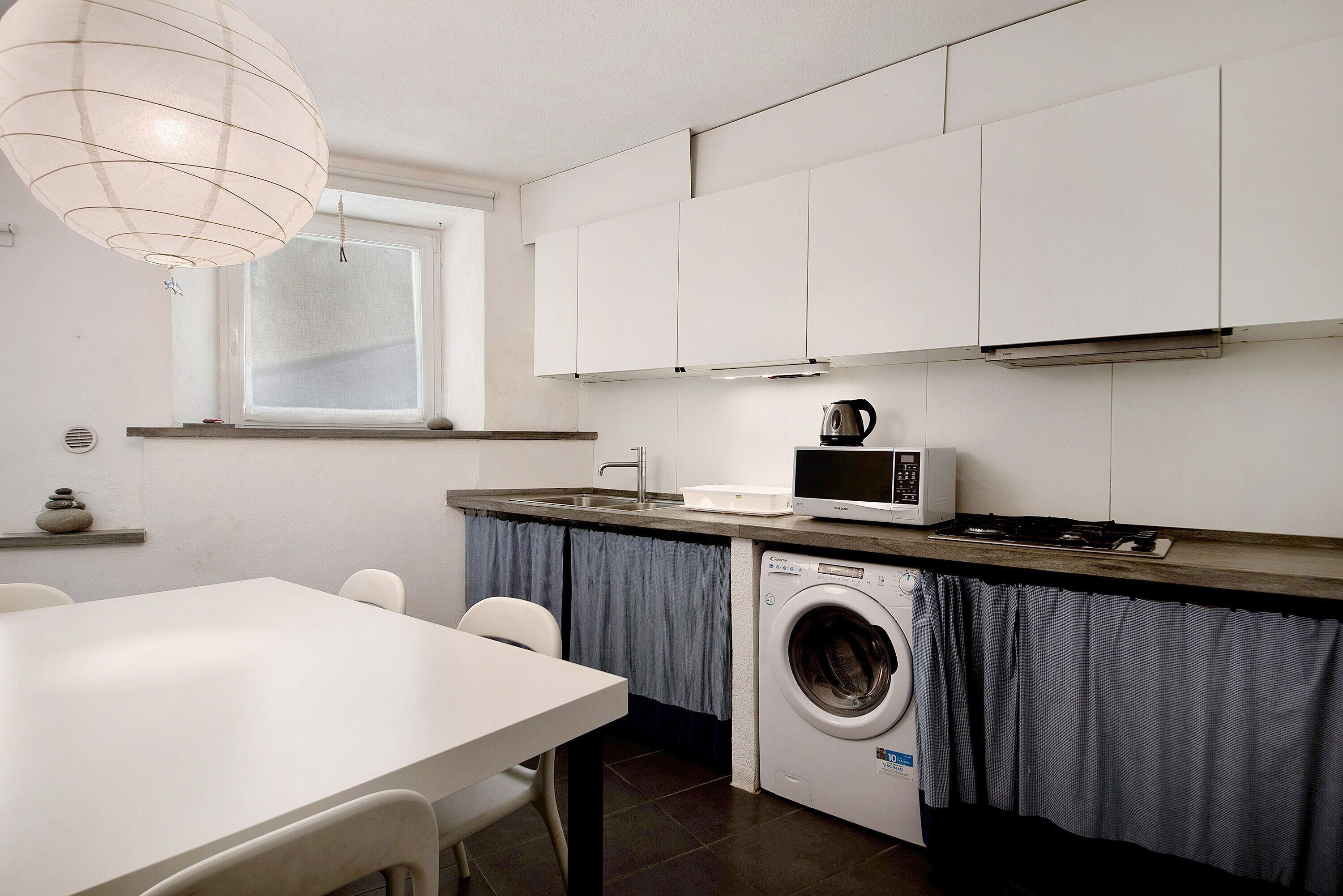 A compact kitchen with a washing machine, a microwave, and a window with curtains.
