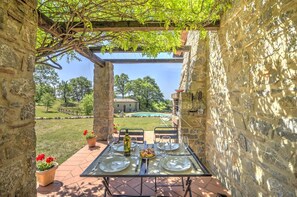 An outdoor dining area with stone walls, a pergola, and a view of a pool and building in the background.