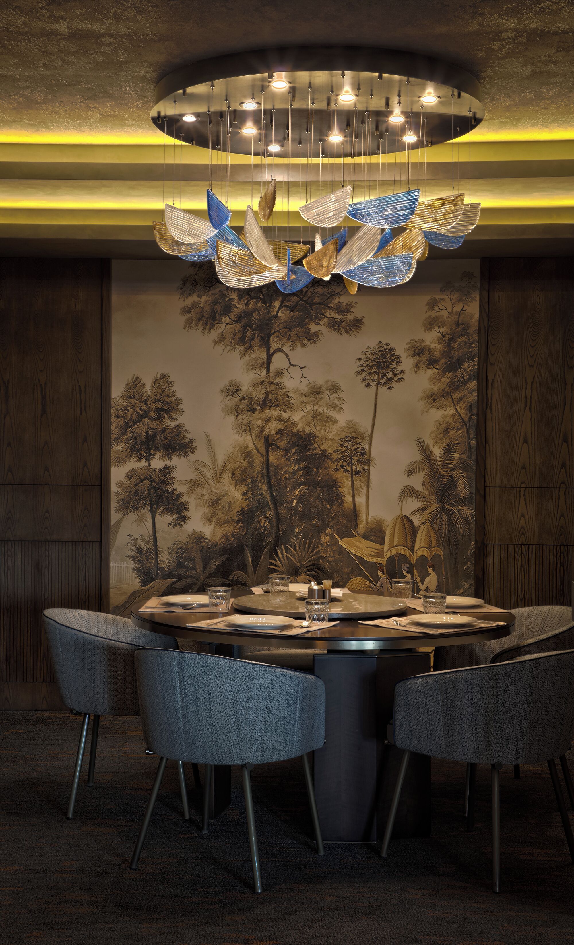 A dining area with a round table, four chairs, a chandelier, and a mural of trees on the wall.