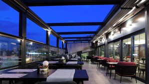 An outdoor dining area with tables and chairs, large windows, and a view of the cityscape. - The Alpfine Hotel (Ankara)