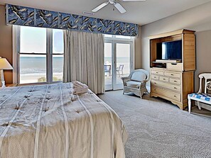 A hotel room with a large bed, a television, a dresser, and a chair. The room has a view of the beach.