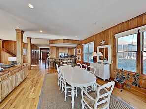A spacious kitchen with wooden paneling, a dining area with white chairs, and large windows offering a view.