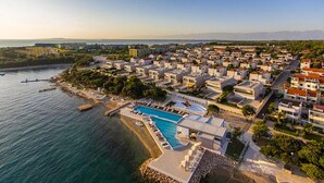 Vista aèria d'una zona residencial costanera amb una gran piscina envoltada de cases modernes, un port esportiu i una vista clara del mar.
