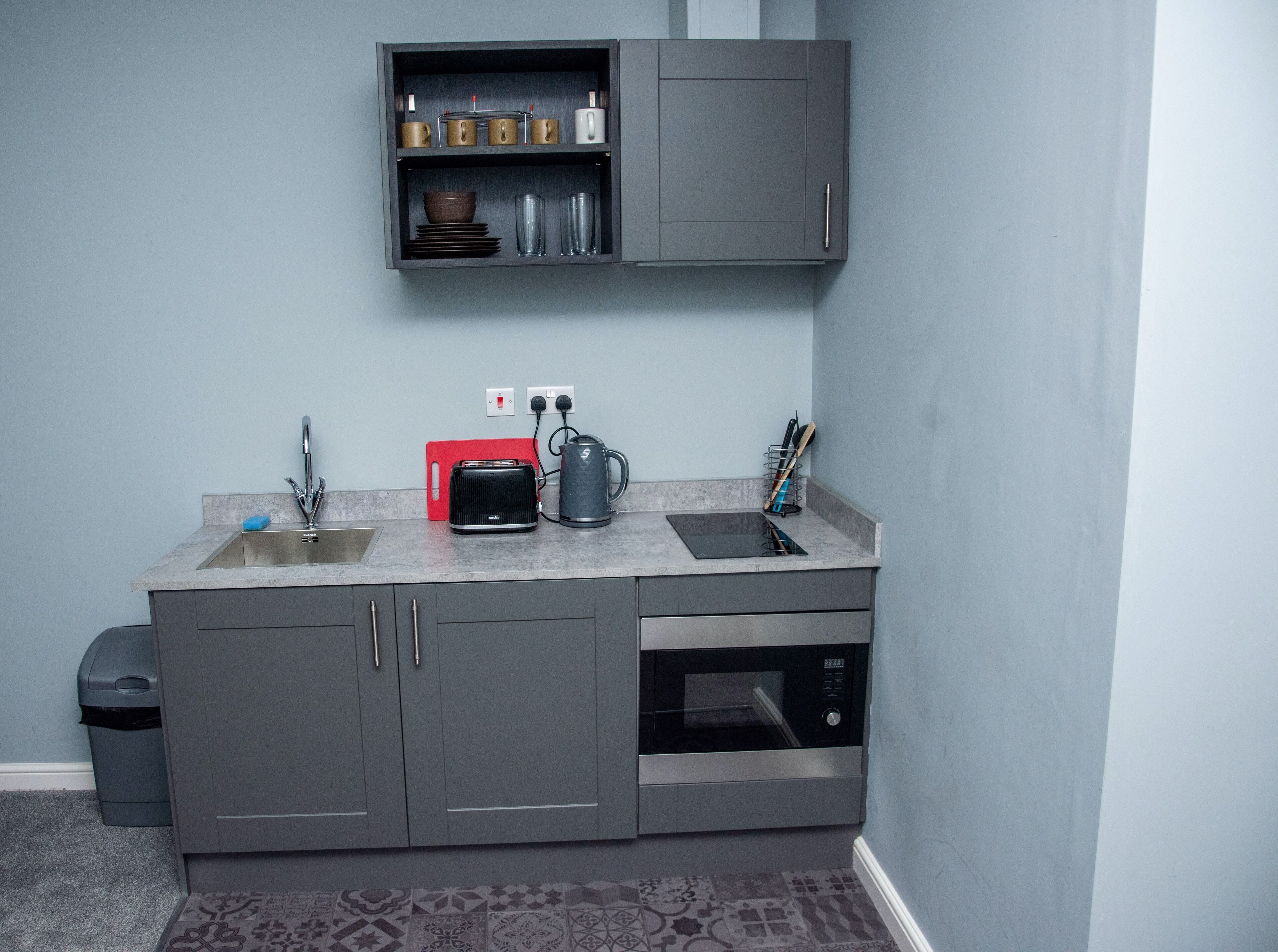 A compact kitchen with grey cabinets, a built-in oven, a toaster, a kettle, and a sink.