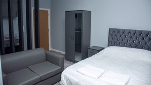 Una habitación de hotel moderna con un sofá tapizado gris, una cama grande con ropa blanca, un armario gris y una mesita de noche pequeña.